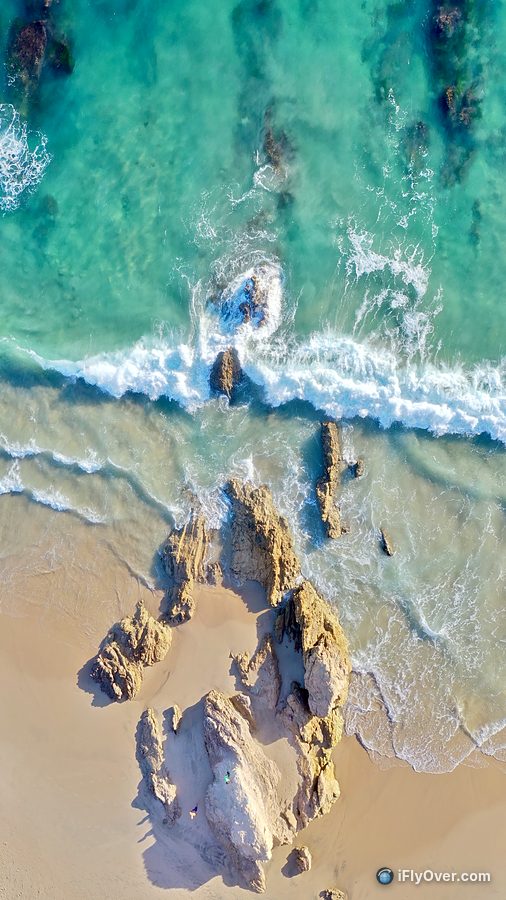 Top-down drone photo of turquoise water and waves washing over rocks on the sand